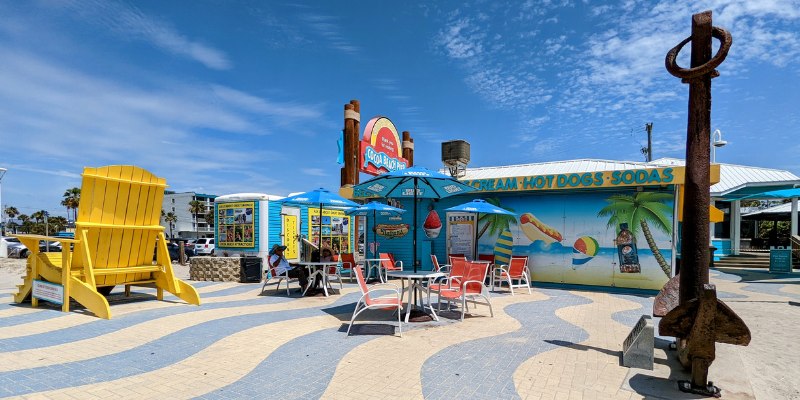 A giant yellow beach chair and a large anchor sit at the entrance of the Cocoa Beach Pier, which features colorful food stands and shops.