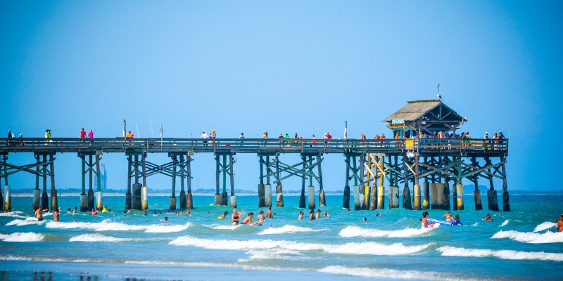 Waves roll into the sandy shore as people swim in the ocean near a long wooden pier, a popular destination accessible from Downtown Cocoa Beach.