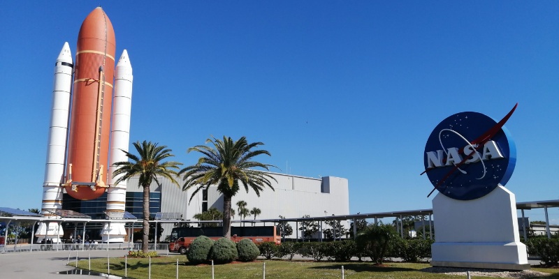 A space shuttle model and a large NASA logo sign stand prominently against a blue sky at the Kennedy Space Center visitor complex.