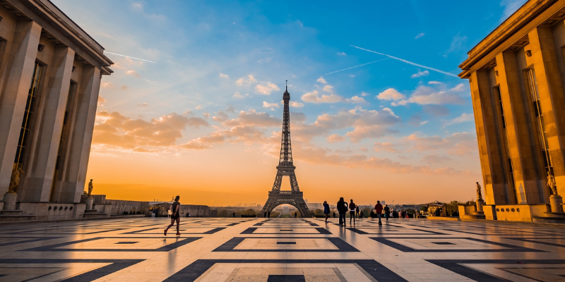 The Eiffel Tower stands tall at sunset, surrounded by a vibrant orange and blue sky as people stroll nearby, enjoying the view.