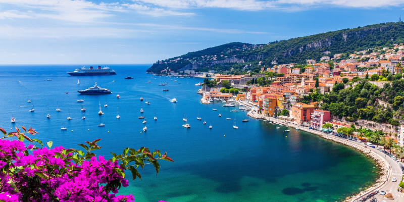 The scenic view of the French Riviera coastline features sailboats and cruise ships, vibrant flowers, and the colorful town of Villefranche-sur-Mer.