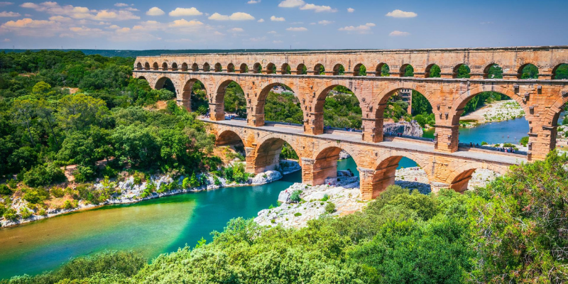 Ancient Roman aqueduct bridge, Pont du Gard, surrounded by lush greenery and flowing river under a blue sky in France.