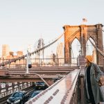 A woman in a stylish coat and hat poses on the Brooklyn Bridge, showcasing the iconic structure and cityscape behind her.