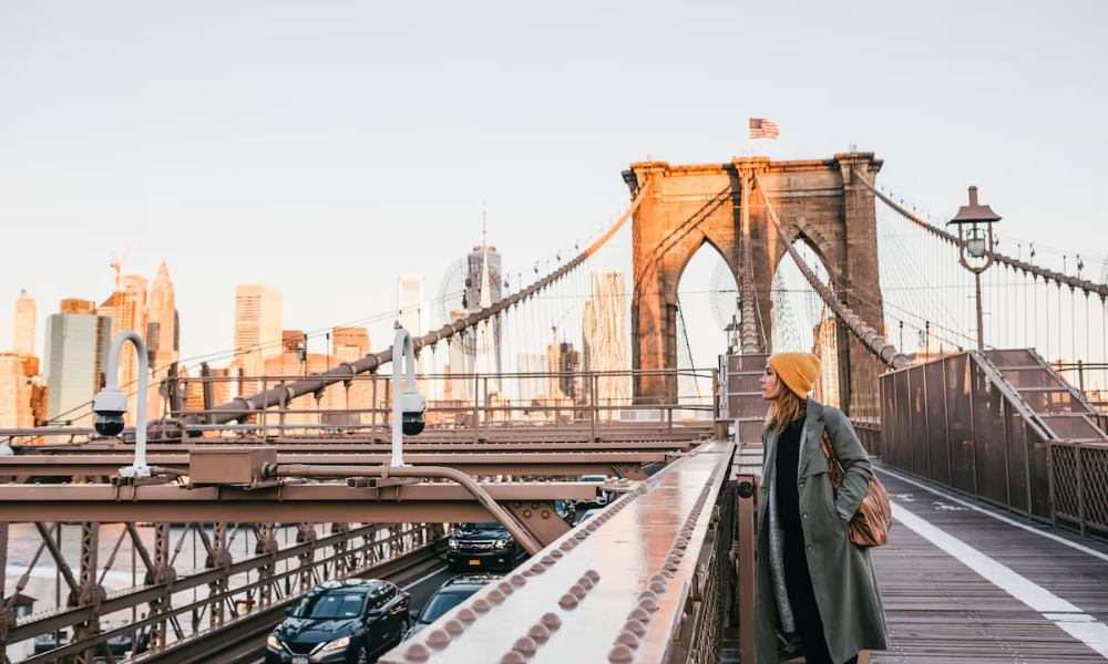 A woman in a stylish coat and hat poses on the Brooklyn Bridge, showcasing the iconic structure and cityscape behind her.