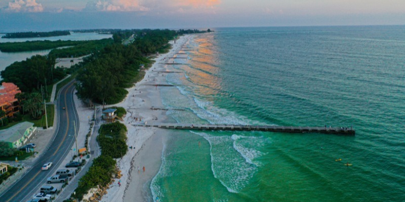 Aerial view of Anna Maria Island featuring a scenic beach with a long pier extending into the turquoise ocean, bordered by lush greenery and a winding coastal road.