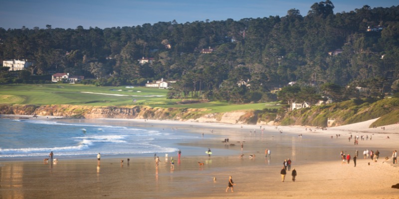 People walk on Carmel Beach, enjoying the sun, with a scenic golf course nearby.