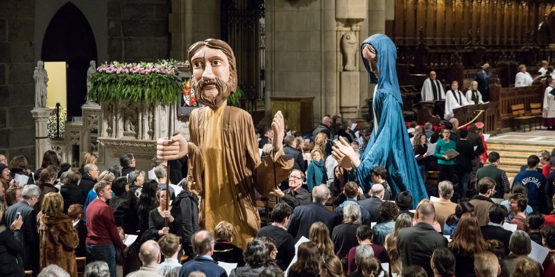 Inside the Cathedral of St. John the Divine, a large congregation is seen with a statue of Jesus prominently displayed.