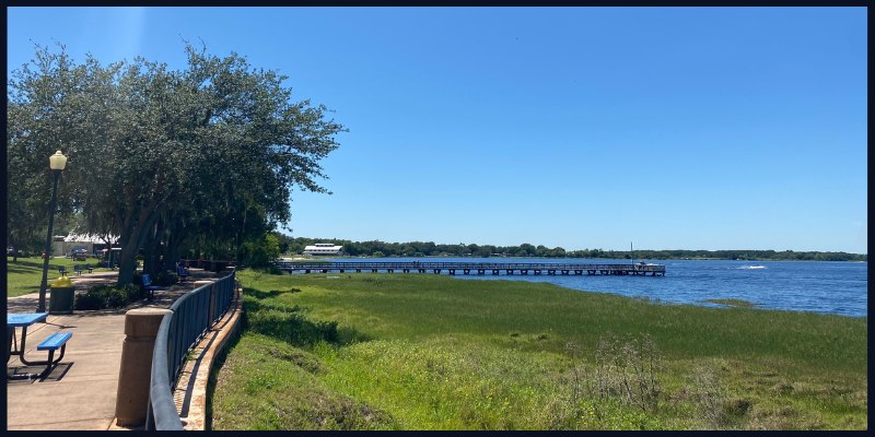 Scenic view of a lakeside park in Clermont, Florida with a walking path, greenery, and a pier extending into the water under a clear blue sky.