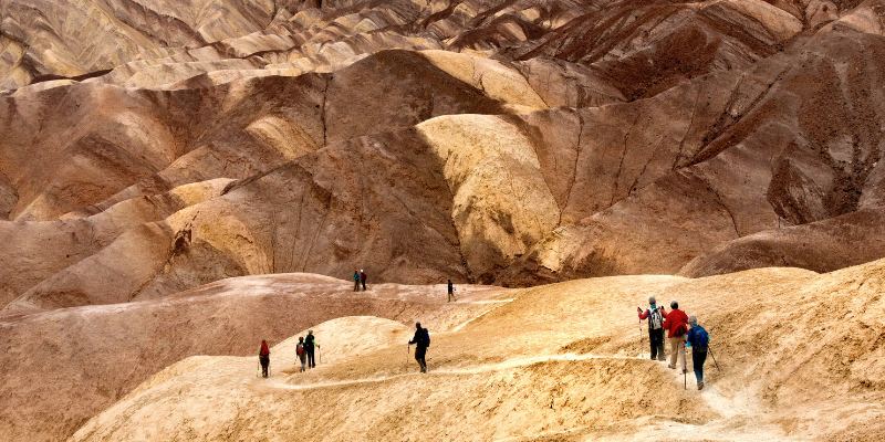 Hikers stroll along a dusty trail in Death Valley National Park, surrounded by vast desert landscapes and clear skies.