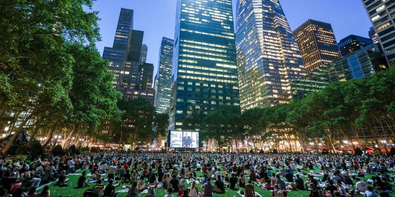 People relax on the grass in Bryant Park, enjoying movies with tall buildings in the background.