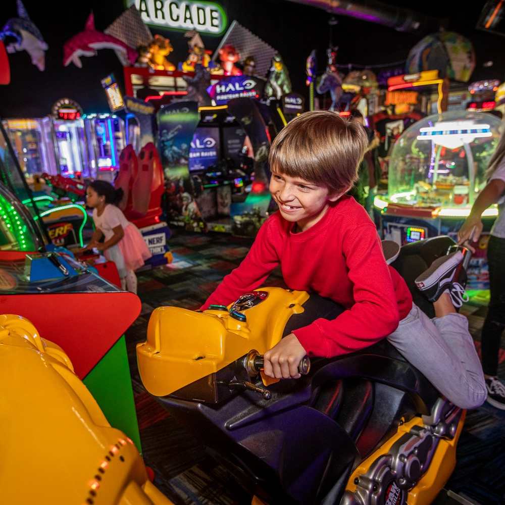 A young boy joyfully rides a motorcycle in an arcade, surrounded by colorful games and bright lights at Fast Eddie's Fun Center.