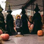 A child in a costume surrounded by people dressed as witches, standing among pumpkins and straw bales, creating a Halloween atmosphere.