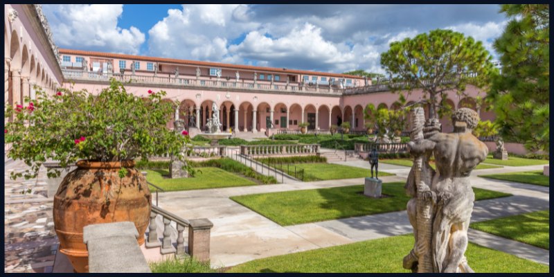 The courtyard of the John and Mable Ringling Museum of Art features classical sculptures, manicured lawns, and a grand colonnade under a blue sky.