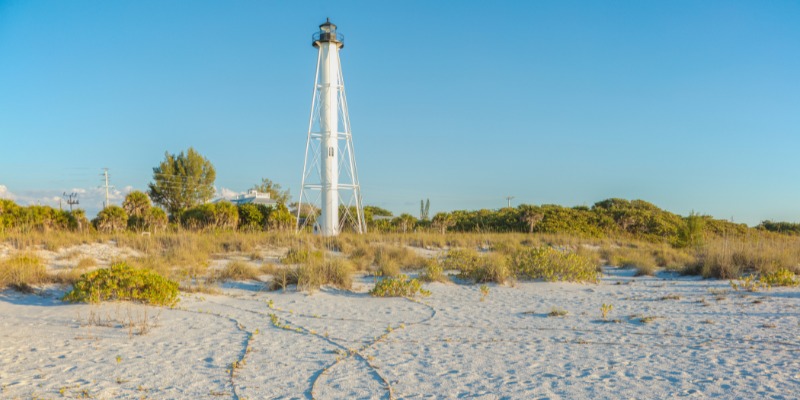 A beautiful white lighthouse is surrounded by sandy dunes and green plants on Little Gasparilla Island, Florida, with a clear blue sky above.