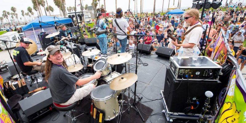 A band performs live outdoors, featuring a smiling drummer and a lively crowd with palm trees in the background.