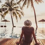 A man in a hat sits by the pool, gazing out at the serene ocean view.