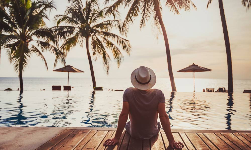 A man in a hat sits by the pool, gazing out at the serene ocean view.