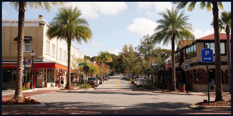 A picturesque street in Mount Dora, Florida, lined with palm trees, charming shops, and cafes under a sunny sky.