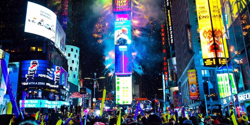 Festive atmosphere in Times Square on New Year's Eve, with a large crowd and iconic ball drop illuminated by colorful lights.