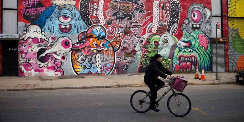 A man cycles by a vibrant monster mural on a building in Bushwick, showcasing the area's dynamic street art scene.