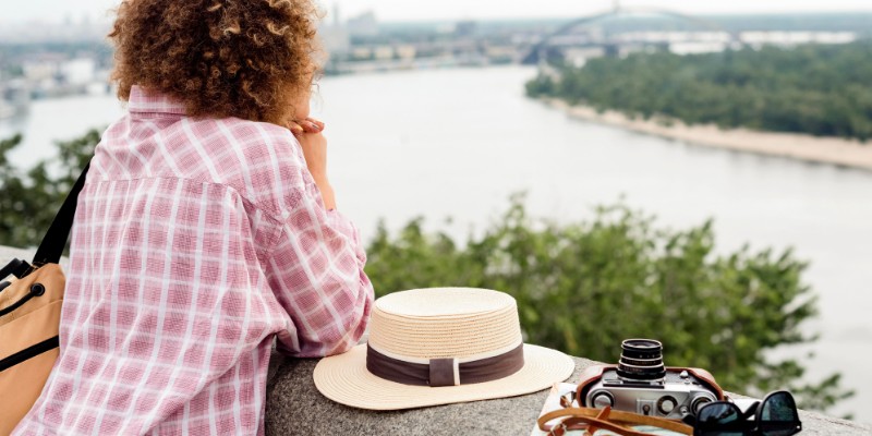 A woman wearing a hat and holding a camera looks over a river, representing travel advice for U.S. elections.