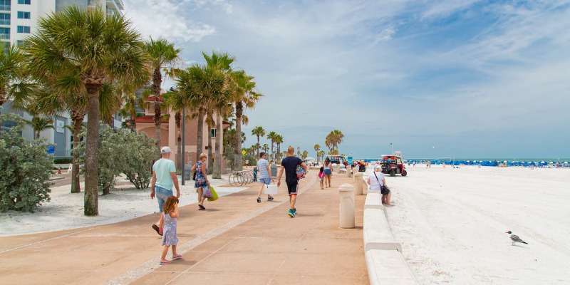 Beachgoers walk on the sand, surrounded by palm trees and buildings under a clear blue sky.