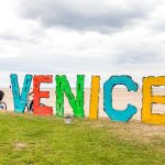 Colorful Venice sign on the beach with people biking nearby on a cloudy day, Venice Beach, California.