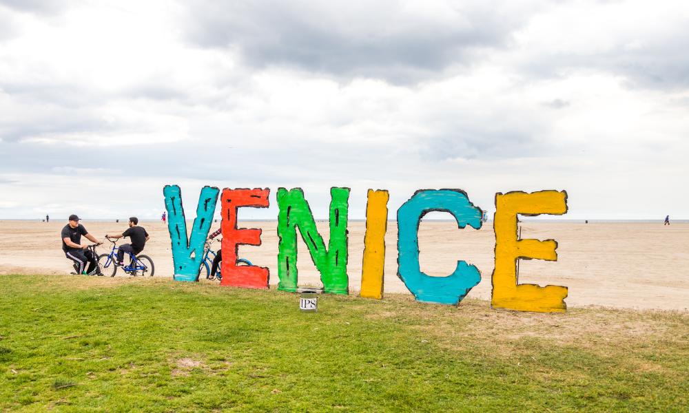 Colorful Venice sign on the beach with people biking nearby on a cloudy day, Venice Beach, California.