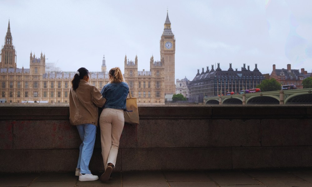 Two women relax on a wall, enjoying a view of the famous Big Ben clock tower nearby.