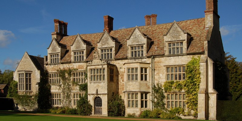 The image depicts Anglesey Abbey, showcasing a substantial stone building adorned with multiple windows and a grassy lawn.