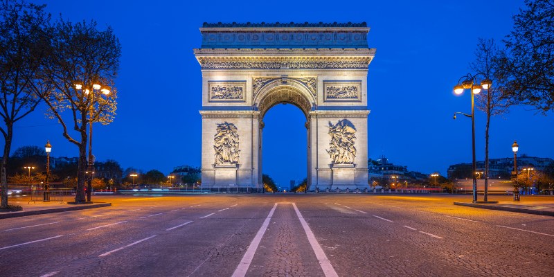 The Arc de Triomphe in Paris shines brightly at night, surrounded by twinkling city lights and a starry sky.