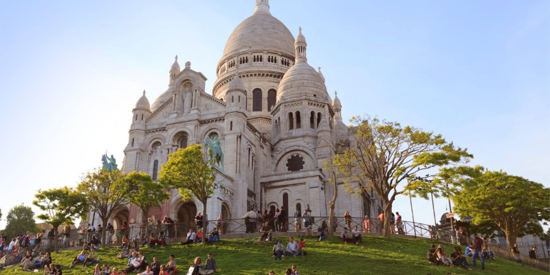 People relax on the lawn in front of the Basilica of Sacré-Cœur on a sunny day.