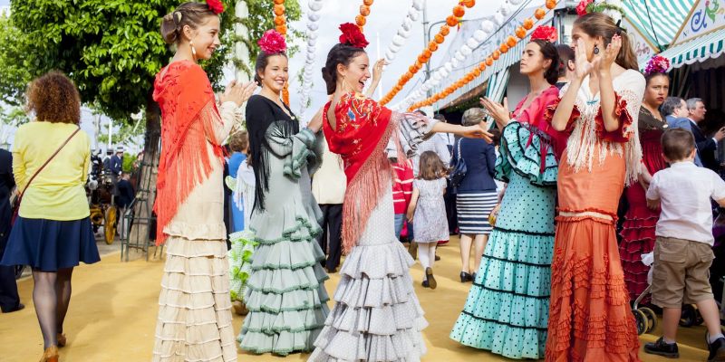 Women in bright, colorful dresses dance together, celebrating the Feria de Abril in Seville.