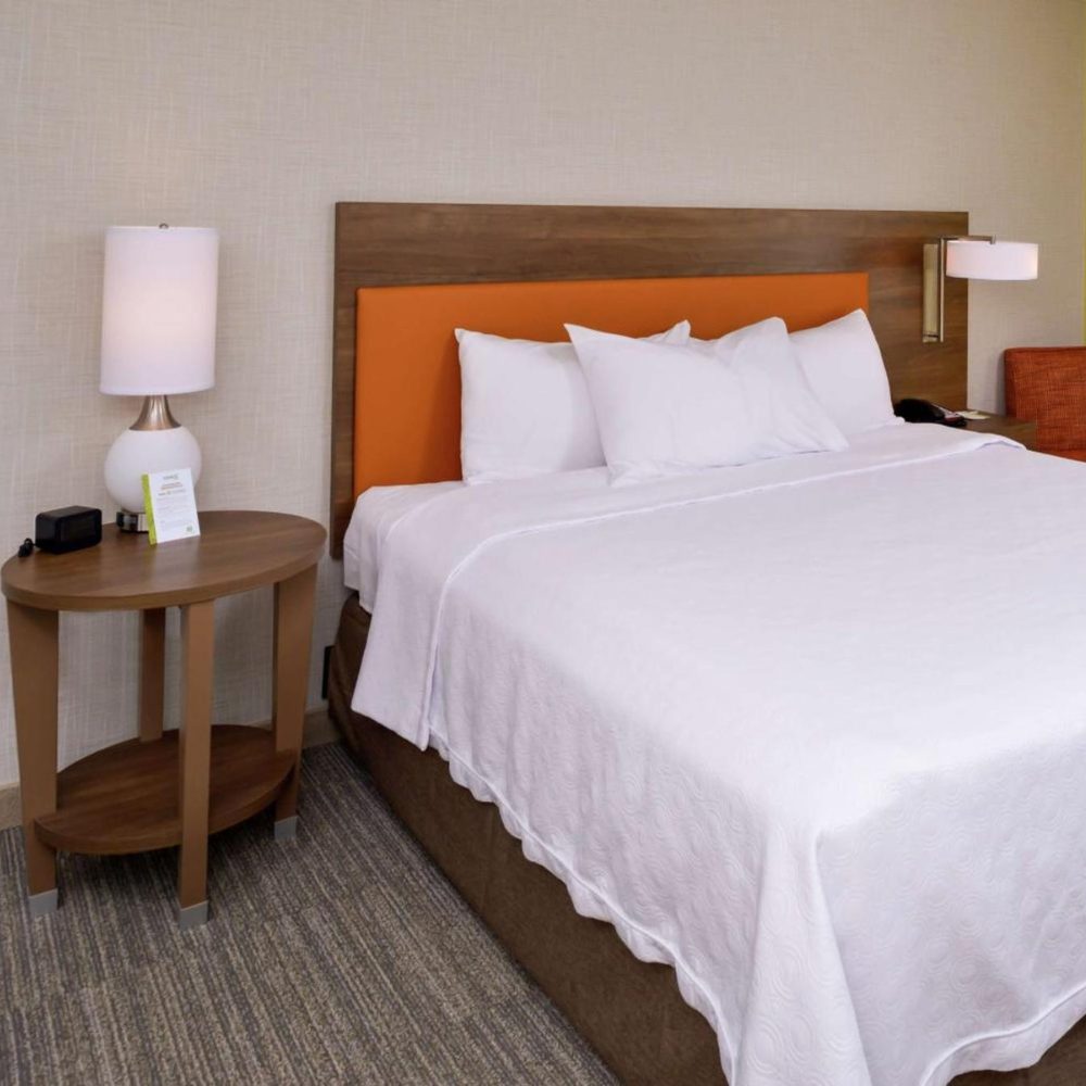 Cozy hotel room at Home2 Suites featuring a bed, nightstand, and a vibrant orange headboard for a cheerful touch.