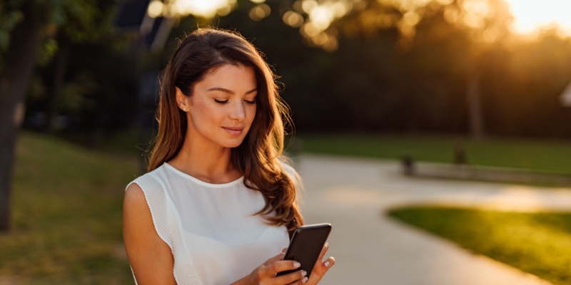 A woman stands in a park, focused on her phone, reminding us to keep emergency contacts handy.