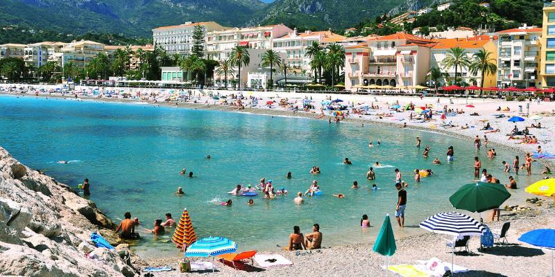 A lively beach in Menton, France, filled with people and umbrellas, creating a festive atmosphere by the water.