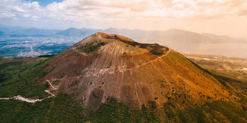 Aerial view of Mount Vesuvius, Italy’s active volcano, with hiking paths and a stunning cityscape and mountain backdrop.