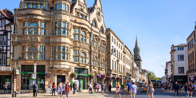 Individuals walk leisurely down a street in Oxford, showcasing a charming building nearby.