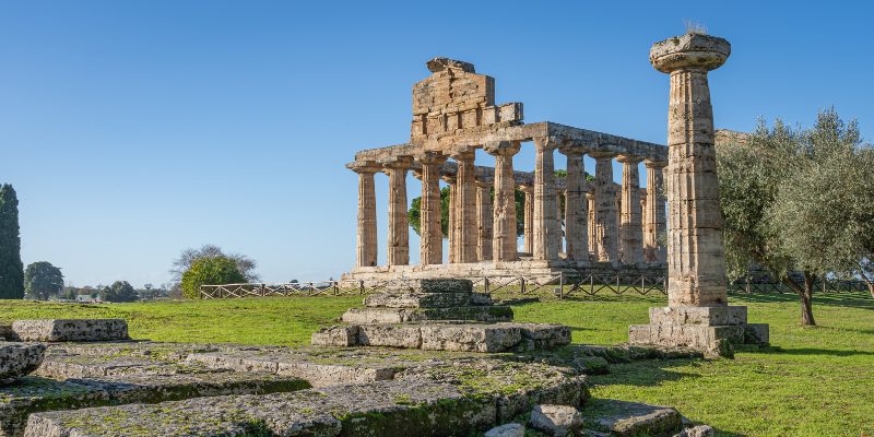 The ancient Greek temple ruins at Paestum in Italy feature stone columns rising majestically under a clear blue sky, surrounded by a lush grassy landscape and whispering trees.