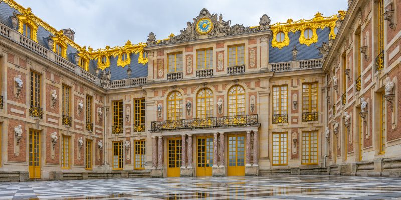 The majestic courtyard of the Palace of Versailles, features intricate architecture and lush greenery.