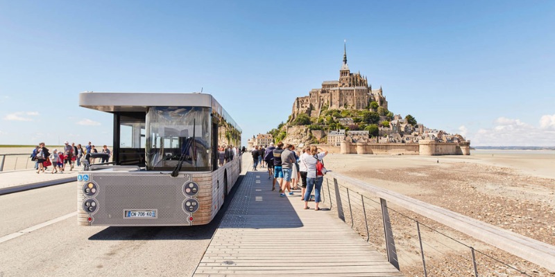 Tour bus traveling along the road towards Mont Saint Michel, with parking and shuttle services visible nearby.