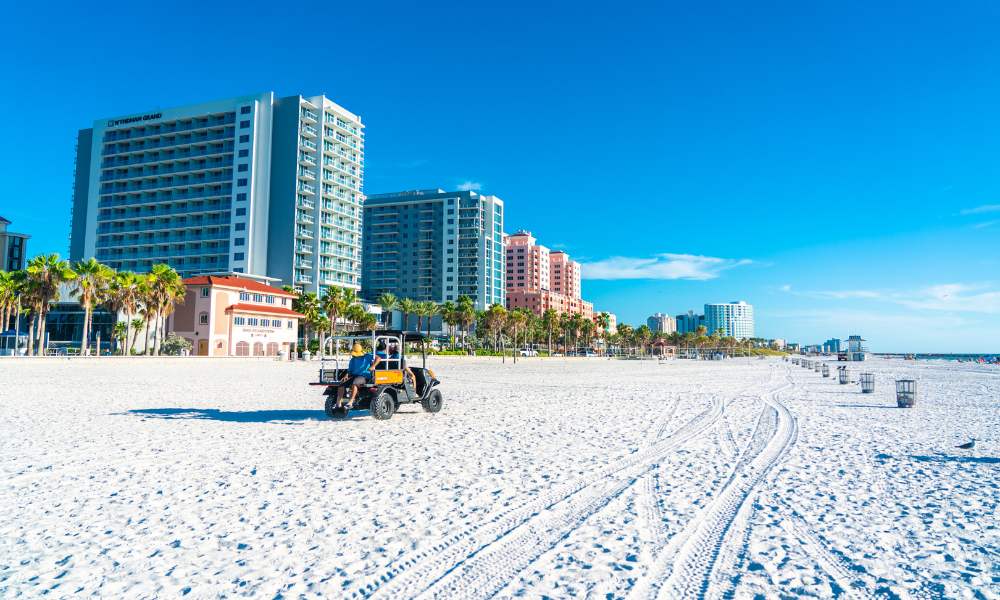 A man in a golf cart navigates Clearwater Beach, tall buildings visible behind him under a bright blue sky.