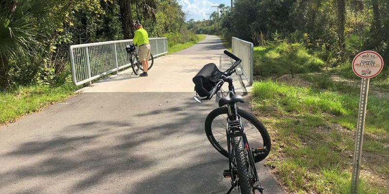 Two bicycles are parked along the Coast-to-Coast Trail in Florida, with trees and a bridge nearby.