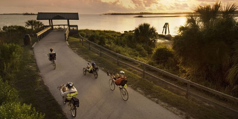 Cyclists ride on the Withlacoochee State Trail in Florida at sunset, with trees and a body of water beside the trail.