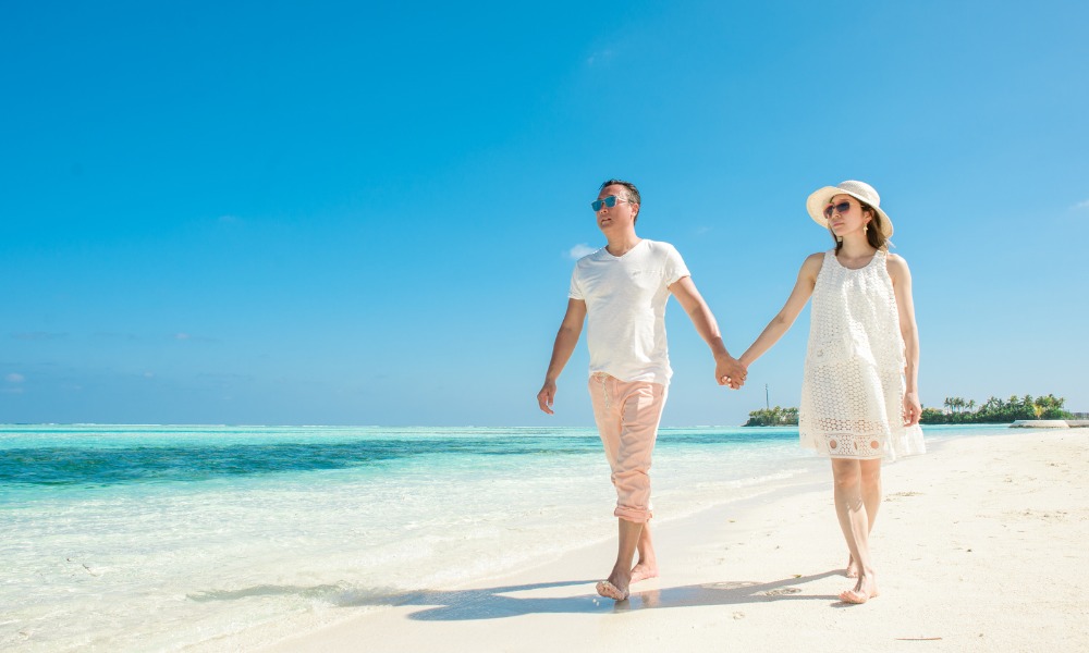 A couple walks together on the beach, hand in hand, with the sun shining and waves lapping at the shore.