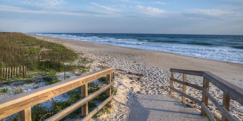 A view of a beach path, showing soft sand and ocean waves under a bright blue sky.