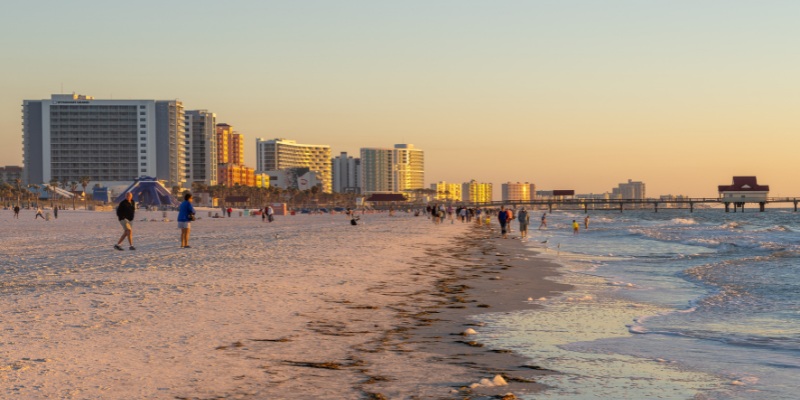 Clearwater Beach features people walking along the shore at sunset, framed by tall buildings in the background.