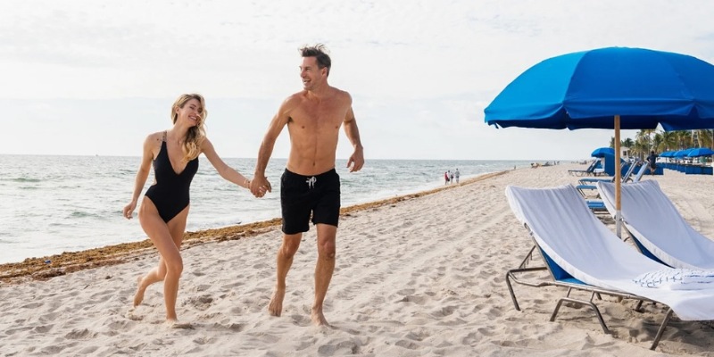 A couple in swimsuits running together on Fort Lauderdale Beach, with the ocean and sky in the background.