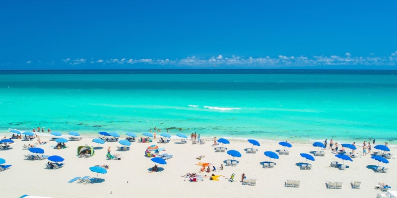 A crowded Miami Beach with numerous people relaxing under colorful umbrellas on the sandy shore.