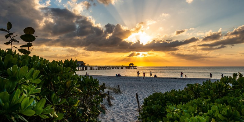 Sunset over Naples Beach with people walking along the sandy shore, casting long shadows in the warm evening light.